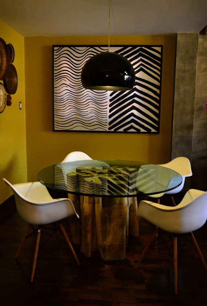 El color de la pared es un amarillo verdoso llamado Chartreuse. Cuadro de tela de origen sueco marca Marimekko diseñada en los años 60. Se usó un vidrio de otro mueble para la actual mesa de comedor, sobre una base de raíz natural de madera de naranjillo.   La lámpara es italiana, marca Kartell. El color negro del policarbonato contrasta con el blanco de las sillas